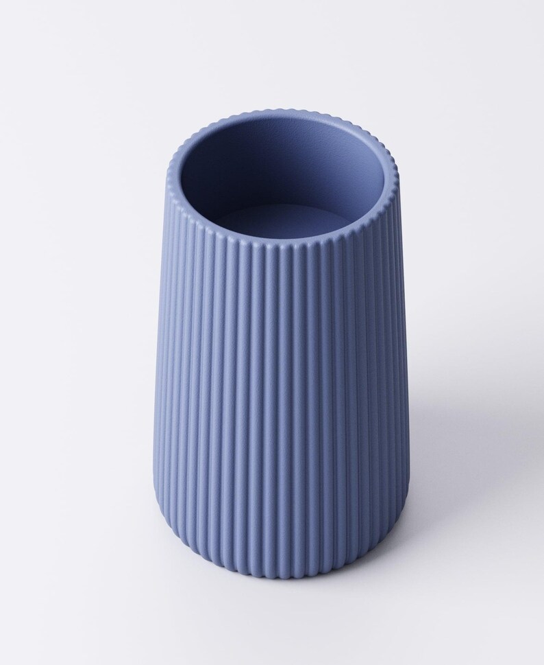 May include: A blue-gray, ribbed vase with a cylindrical shape. The vase has a slightly flared base and a textured surface with vertical lines. The top rim has a decorative, ridged edge. The vase is empty and sits on a white background.