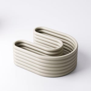 May include: A light grey, modern ceramic decorative bowl. The bowl has a unique, wavy design with multiple horizontal ridges. The neutral colour and minimalist style make it a versatile piece for home decor.