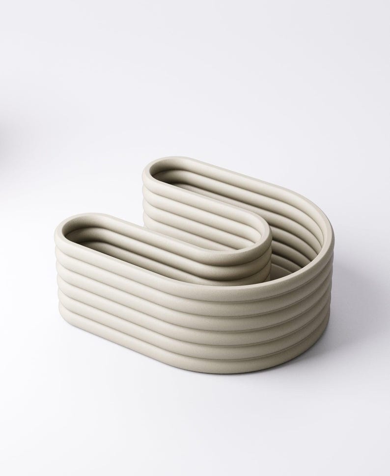 May include: A light grey, modern ceramic decorative bowl. The bowl has a unique, wavy design with multiple horizontal ridges. The neutral colour and minimalist style make it a versatile piece for home decor.