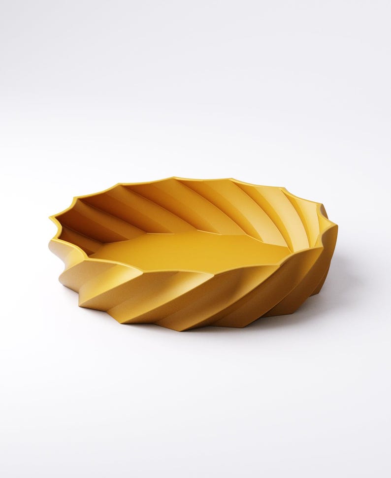 May include: A mustard-yellow decorative bowl with a unique, ridged design. The bowl has a shallow, oval shape with a textured exterior. The ridges create a spiral effect around the bowl's edge. The interior is smooth and the bowl is set against a white background.
