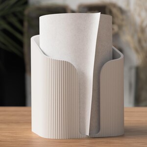 Op de afbeelding: Een witte papieren handdoekhouder met een verticaal geribbeld ontwerp. De houder heeft een gebogen opening om de papieren handdoeken af te geven. Een rol papieren handdoeken bevindt zich in de houder, met een vel gedeeltelijk uitgetrokken.