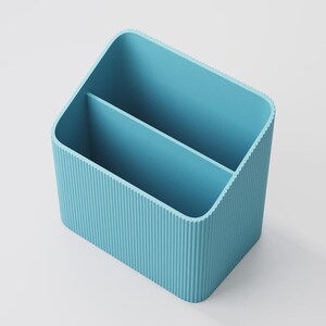 Puede incluir: Un organizador de escritorio azul claro con dos compartimentos. El organizador tiene forma rectangular con esquinas redondeadas y detalles acanalados verticales en los laterales. El interior es liso y el dise&ntilde;o general es minimalista.