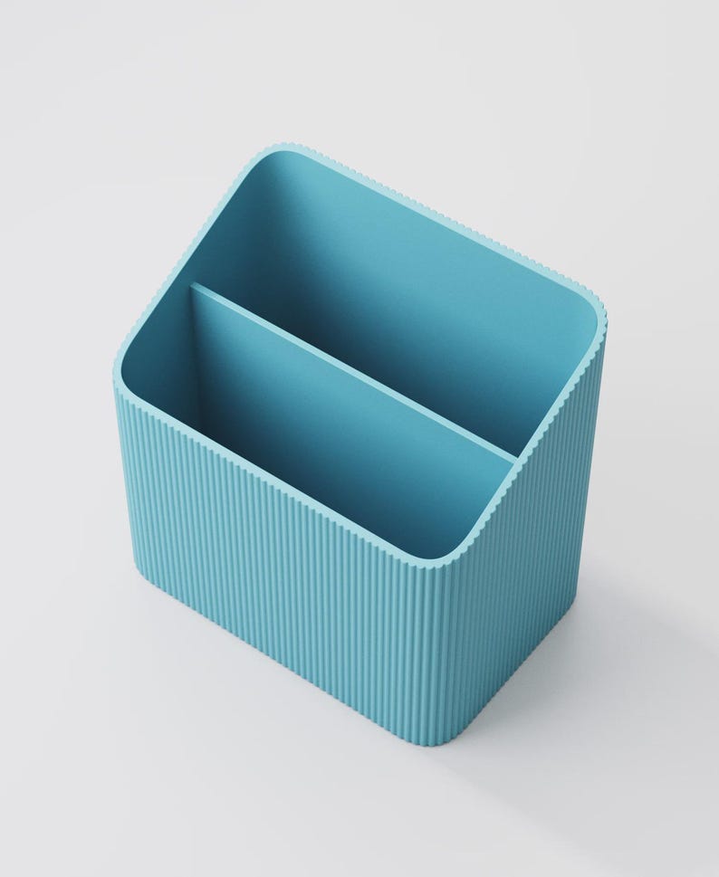 Puede incluir: Un organizador de escritorio azul claro con dos compartimentos. El organizador tiene forma rectangular con esquinas redondeadas y detalles acanalados verticales en los laterales. El interior es liso y el dise&ntilde;o general es minimalista.
