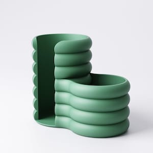 May include: A green, modern desk organizer with a unique, curved design. The organizer features a series of stacked, rounded forms, creating compartments for storage. The color is a muted, earthy green, and the overall aesthetic is minimalist and contemporary.