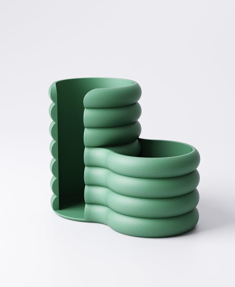 May include: A green, modern desk organizer with a unique, curved design. The organizer features a series of stacked, rounded forms, creating compartments for storage. The color is a muted, earthy green, and the overall aesthetic is minimalist and contemporary.