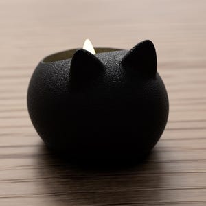 May include: A black cat-shaped candle holder with a textured surface. The candle holder has two pointed ears and a round body. A small, lit candle is inside the holder, emitting a soft glow. The holder is sitting on a wooden surface.