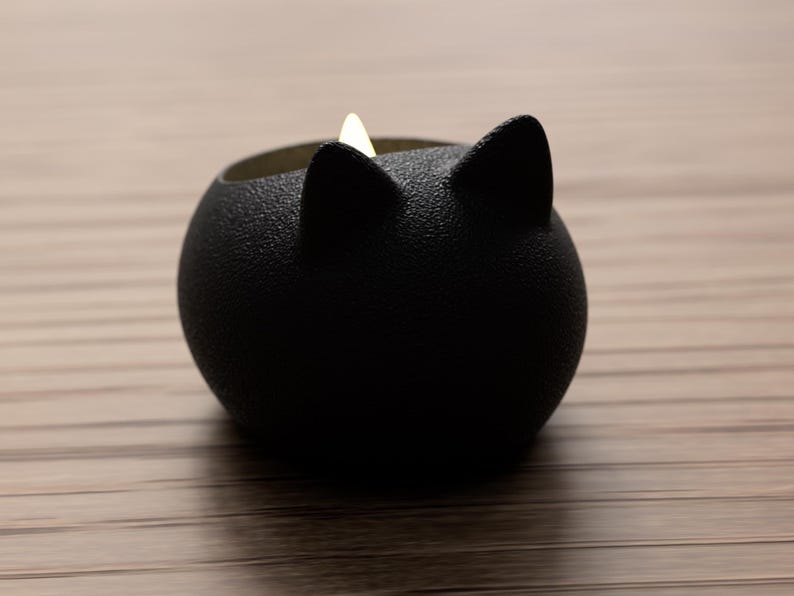 May include: A black cat-shaped candle holder with a textured surface. The candle holder has two pointed ears and a round body. A small, lit candle is inside the holder, emitting a soft glow. The holder is sitting on a wooden surface.