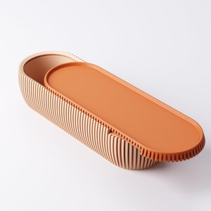 May include: An oval-shaped wooden storage box with a lid. The box is made of vertical wooden slats and has a light brown color. The lid is a solid orange color. The box is open, revealing the interior.