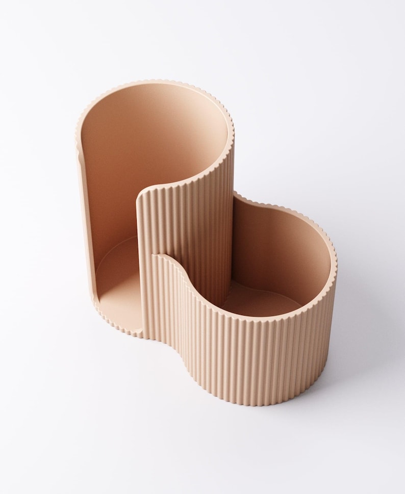 May include: A peach-colored desk organizer with a unique, wavy design. The organizer has three distinct sections: a tall, open compartment, a smaller, curved section, and a wider, open area. The exterior features vertical ribbed detailing.