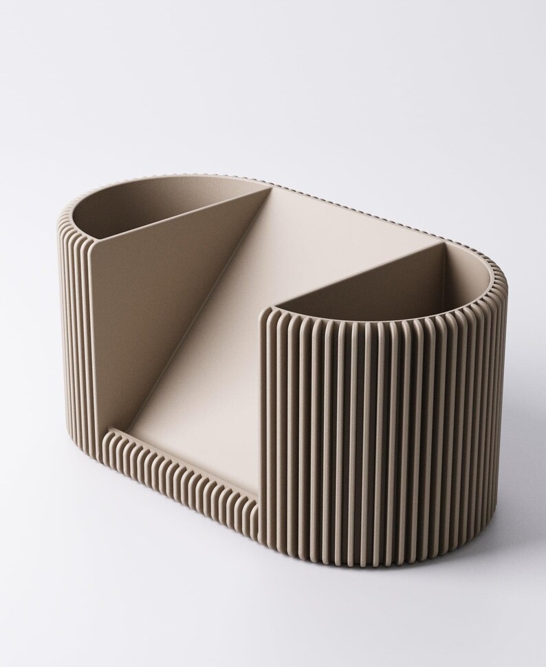 May include: A beige desk organiser with a distinctive design. It features two rounded compartments and a central section, all with vertical ribbed detailing. The oval shape and neutral colour make it suitable for any office or home decor.