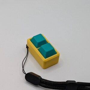 May include: A yellow and teal keycap set with a black wrist strap. The keycaps are teal and square, set in a yellow rectangular frame. The wrist strap is black and braided, with black plastic end pieces.
