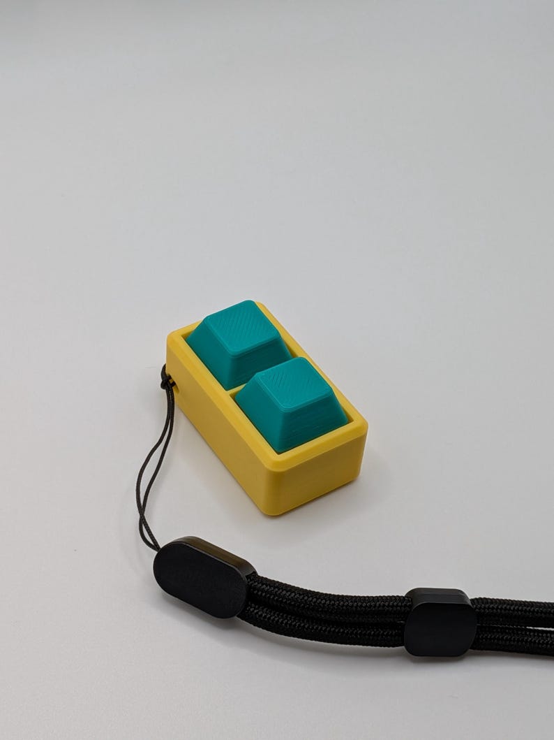 May include: A yellow and teal keycap set with a black wrist strap. The keycaps are teal and square, set in a yellow rectangular frame. The wrist strap is black and braided, with black plastic end pieces.