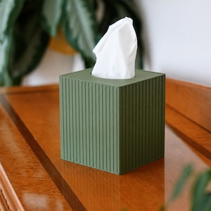 May include: A square olive green tissue box cover featuring a vertical ribbed texture on all four sides. A single white tissue is pulled through the square opening at the top of the container, which is designed to hold standard square tissue boxes.