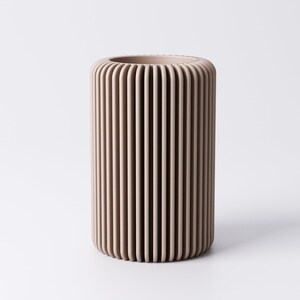 May include: A cylindrical vase in a neutral beige color. The vase has a ribbed design with vertical lines running from top to bottom. The vase is made of a solid material and has a smooth, matte finish. The vase is suitable for home decor.