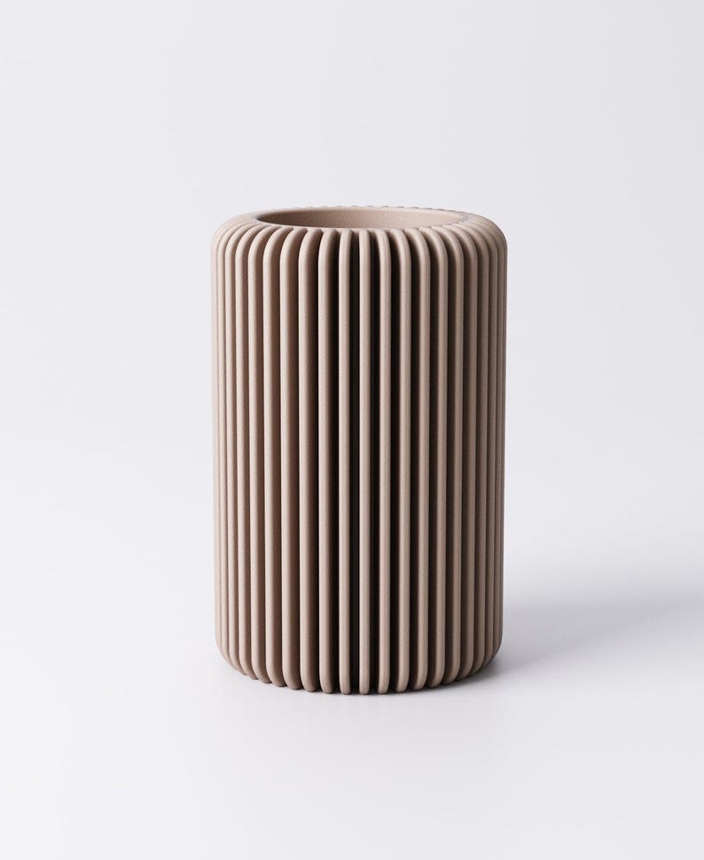 May include: A cylindrical vase in a neutral beige color. The vase has a ribbed design with vertical lines running from top to bottom. The vase is made of a solid material and has a smooth, matte finish. The vase is suitable for home decor.