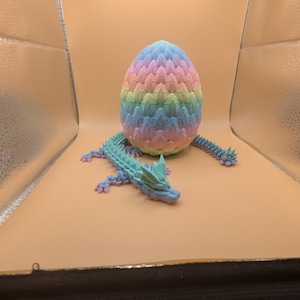 May include: A 3D-printed dragon and egg set. The egg is a pastel rainbow of pink, yellow, blue, and green. The dragon is a matching blue-green with a spiky tail and head. The set is designed to be a decorative item.