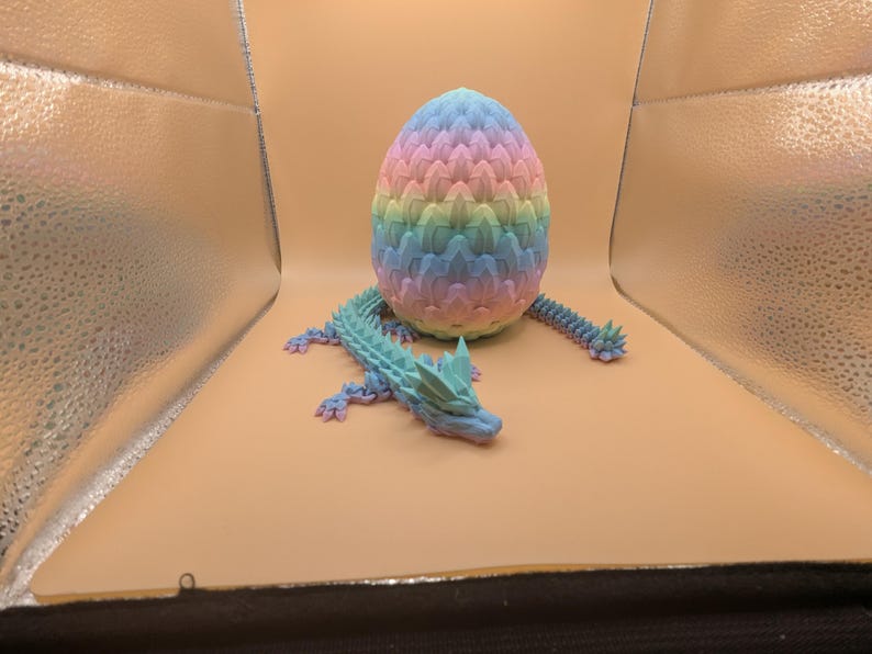 May include: A 3D-printed dragon and egg set. The egg is a pastel rainbow of pink, yellow, blue, and green. The dragon is a matching blue-green with a spiky tail and head. The set is designed to be a decorative item.