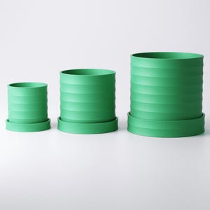 Op de afbeelding: Drie groene cilindrische bloempotten van verschillende formaten, elk met een bijpassende schotel. De potten hebben een geribbeld ontwerp aan de buitenkant. De kleinste pot is ongeveer 10 cm hoog, de middelste 15 cm en de grootste 20 cm.