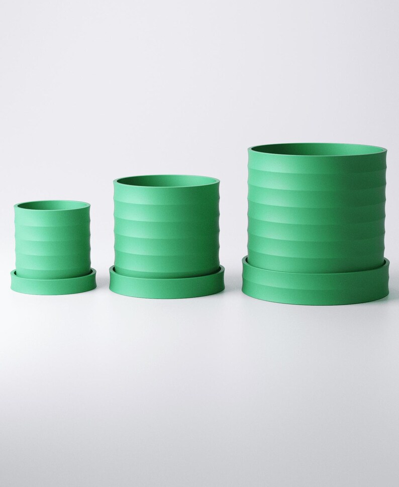 Op de afbeelding: Drie groene cilindrische bloempotten van verschillende formaten, elk met een bijpassende schotel. De potten hebben een geribbeld ontwerp aan de buitenkant. De kleinste pot is ongeveer 10 cm hoog, de middelste 15 cm en de grootste 20 cm.