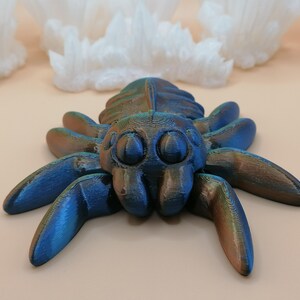 Op de afbeelding: Een 3D-geprinte spinnenfiguur met een metallic blauwe en bronzen afwerking. De spin heeft grote, ronde ogen en acht poten. De achtergrond toont witte, kristallijne formaties tegen een lichtbeige oppervlak.