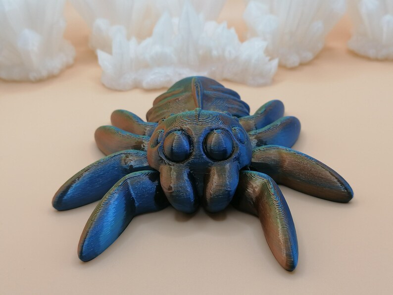 Op de afbeelding: Een 3D-geprinte spinnenfiguur met een metallic blauwe en bronzen afwerking. De spin heeft grote, ronde ogen en acht poten. De achtergrond toont witte, kristallijne formaties tegen een lichtbeige oppervlak.