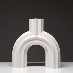 May include: A white, abstract sculpture with an arched design. The sculpture has a vertical, rectangular top section and a rounded, arch-like base. The piece is made of a solid, opaque material and is set against a dark background.