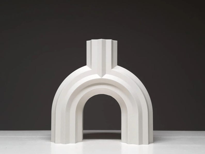 May include: A white, abstract sculpture with an arched design. The sculpture has a vertical, rectangular top section and a rounded, arch-like base. The piece is made of a solid, opaque material and is set against a dark background.