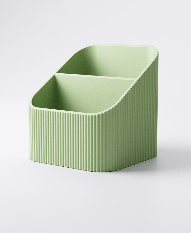 May include: A light green desk organizer with two compartments. The organizer has a ribbed texture on the exterior and a curved top. The item is designed to hold pens, pencils, or other office supplies.