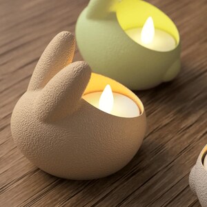 K&ouml;nnte beinhalten: Nahaufnahme von drei hasenf&ouml;rmigen Kerzenhaltern in Beige und Gr&uuml;n, die jeweils eine brennende wei&szlig;e Kerze halten. Die Kerzenhalter stehen auf einer Holzoberfl&auml;che. Die Hasenohren sind erhoben, und die Kerzenflammen leuchten hell.