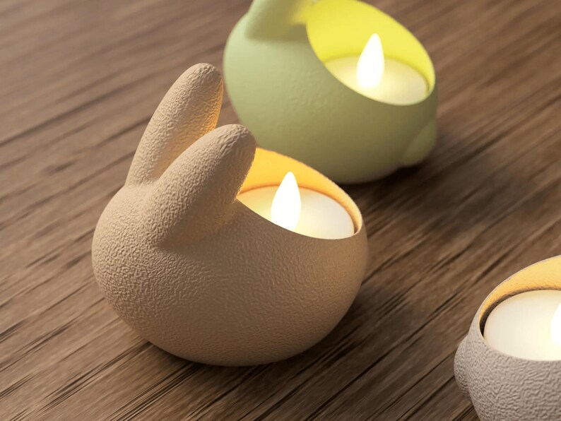 K&ouml;nnte beinhalten: Nahaufnahme von drei hasenf&ouml;rmigen Kerzenhaltern in Beige und Gr&uuml;n, die jeweils eine brennende wei&szlig;e Kerze halten. Die Kerzenhalter stehen auf einer Holzoberfl&auml;che. Die Hasenohren sind erhoben, und die Kerzenflammen leuchten hell.