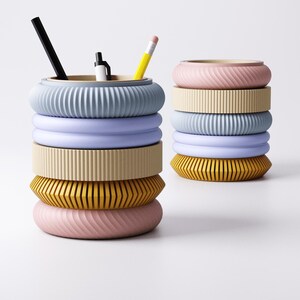 K&ouml;nnte beinhalten: Zwei Stapel bunter Schreibtisch-Organizer. Ein Stapel hat eine hellblaue Oberseite mit geripptem Design, die einen schwarzen Stift, einen wei&szlig;-schwarzen Marker und einen gelben Bleistift enth&auml;lt. Der andere Stapel hat eine rosa Oberseite mit einem Wirbeldesign.