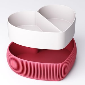 May include: A two-piece heart-shaped storage container. The top section is white and divided into two compartments. The bottom section is a ribbed, deep pink. The container is designed to be stacked, with the top section sitting on the bottom.