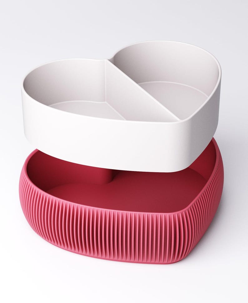 May include: A two-piece heart-shaped storage container. The top section is white and divided into two compartments. The bottom section is a ribbed, deep pink. The container is designed to be stacked, with the top section sitting on the bottom.