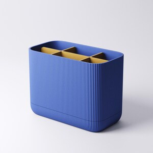 May include: A rectangular desk organizer with a vibrant blue exterior featuring a vertical ribbed texture. The interior is divided into six compartments by mustard yellow dividers, creating a structured space for holding various small items.