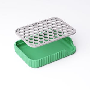 May include: A green rectangular container with a gray, honeycomb-patterned lid. The container has a ribbed exterior and a smooth interior. The lid is slightly elevated above the container, suggesting it is removable.