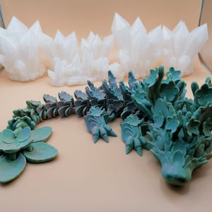 May include: A collection of 3D-printed decorative items. The image features several white crystal formations and a teal-colored, articulated dragon with floral details. A teal-colored succulent is also present. The items are arranged on a light tan surface.