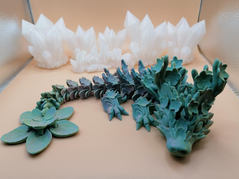 May include: A collection of 3D-printed decorative items. The image features several white crystal formations and a teal-colored, articulated dragon with floral details. A teal-colored succulent is also present. The items are arranged on a light tan surface.