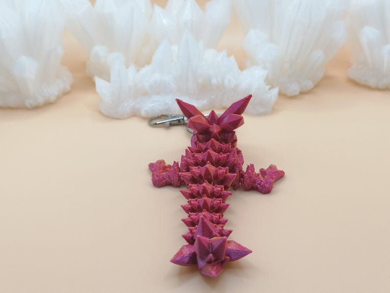 Może przedstawiać: Brelok w kształcie kryształu w kolorze magenta ze srebrnym zapięciem. Kryształ ma ostre, spiczaste krawędzie i teksturowaną powierzchnię. Białe, chmuropodobne formacje w tle kontrastują z magentowym brelokiem.