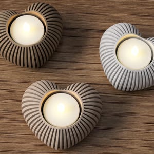May include: Three heart-shaped candle holders with a ribbed design, each holding a lit tea light. The candle holders are in shades of gray and brown, set on a wooden surface. A decorative item for home decor.