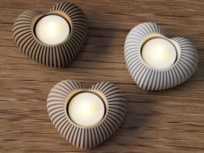 May include: Three heart-shaped candle holders with a ribbed design, each holding a lit tea light. The candle holders are in shades of gray and brown, set on a wooden surface. A decorative item for home decor.
