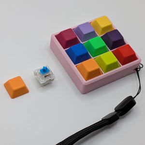 May include: A collection of colorful keyboard keycaps in a pink tray, with a single orange keycap and a clear switch. A black lanyard is also present. The keycaps are various colors, including red, blue, green, and yellow.