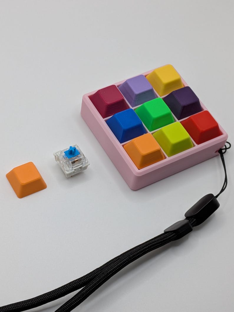 May include: A collection of colorful keyboard keycaps in a pink tray, with a single orange keycap and a clear switch. A black lanyard is also present. The keycaps are various colors, including red, blue, green, and yellow.