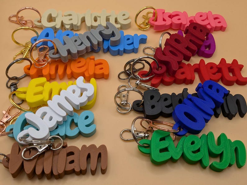 May include: A collection of colorful name keychains in various colors, including pink, blue, and green. Each keychain features a different name in a bold, 3D font, attached to a metal keyring. The names include Charlotte, Henry, and Evelyn.
