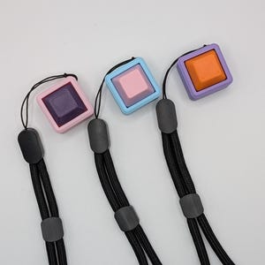 May include: Three square fidget toys in pink, blue, and purple with black lanyards. Each square has a different colored center button. The pink square has a purple button, the blue square has a pink button, and the purple square has an orange button.