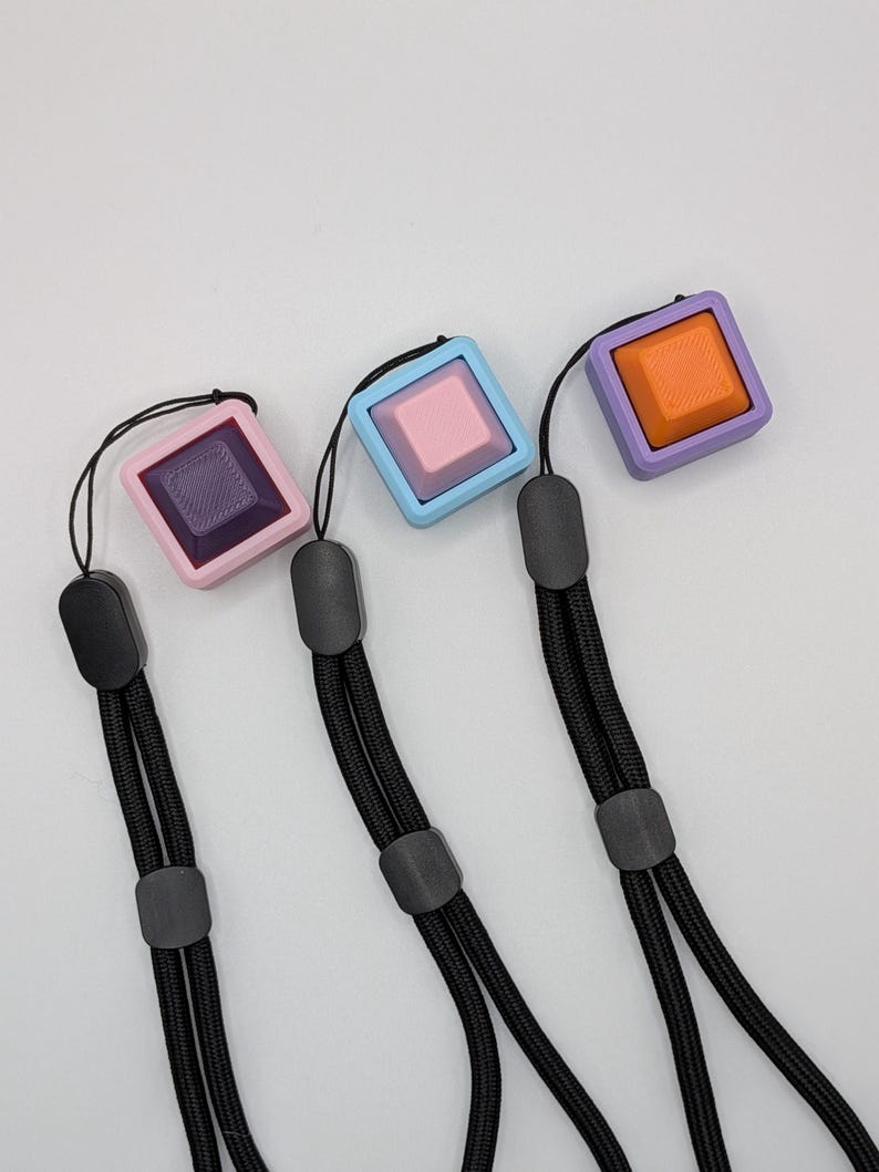 May include: Three square fidget toys in pink, blue, and purple with black lanyards. Each square has a different colored center button. The pink square has a purple button, the blue square has a pink button, and the purple square has an orange button.