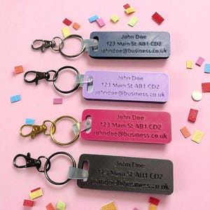 May include: Four rectangular keychains in black, purple, pink, and gray, each with a metal ring and clasp. The keychains display the name "John Doe", an address, and an email address. Colorful confetti is scattered around.