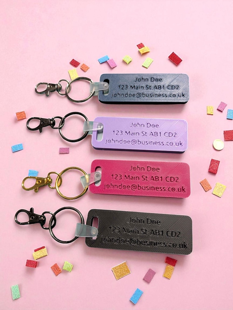 May include: Four rectangular keychains in black, purple, pink, and gray, each with a metal ring and clasp. The keychains display the name "John Doe", an address, and an email address. Colorful confetti is scattered around.