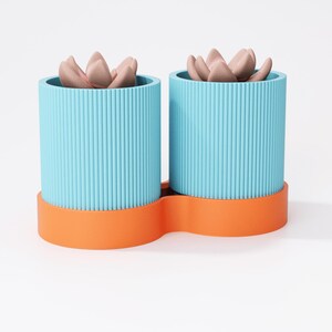 May include: Two blue, ribbed cylindrical planters with orange bases, each holding a faux succulent with pinkish-brown petals. The planters are joined by the orange base, creating a modern design for home decor.
