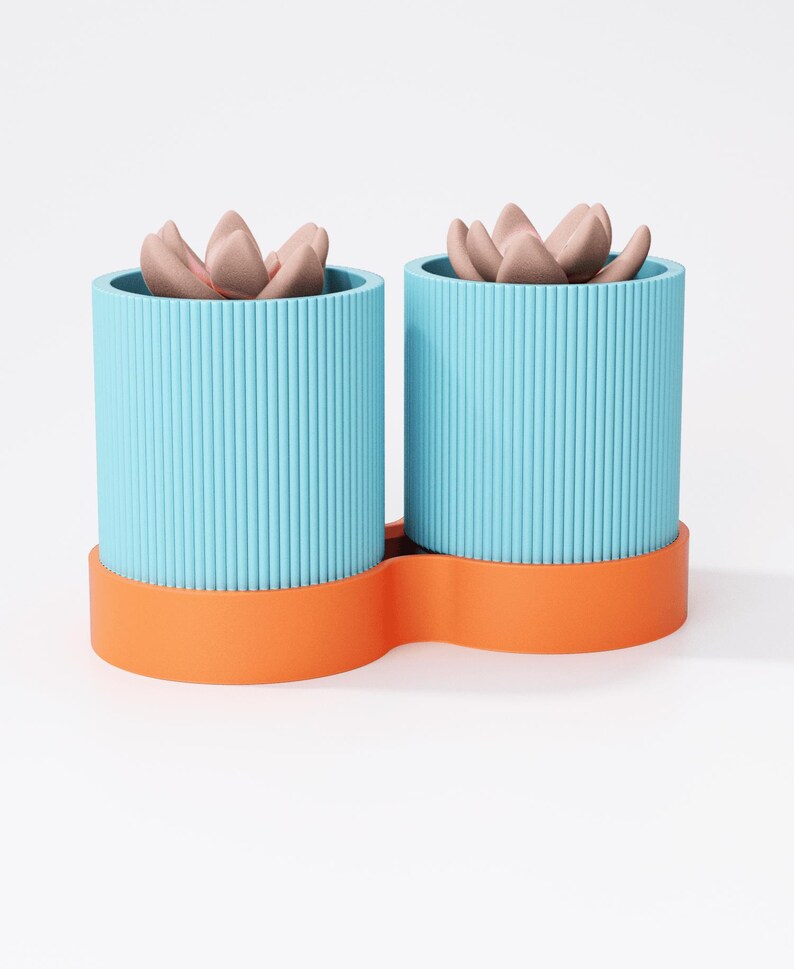 May include: Two blue, ribbed cylindrical planters with orange bases, each holding a faux succulent with pinkish-brown petals. The planters are joined by the orange base, creating a modern design for home decor.
