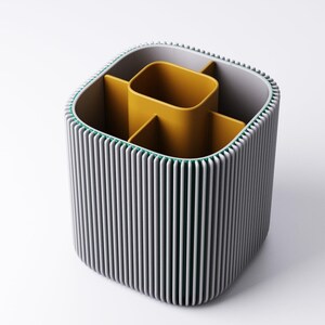 May include: A grey and yellow desk tidy with a ribbed exterior. The organiser has a square shape with rounded corners and a green trim. The interior is divided into compartments, with a central yellow section and additional dividers.